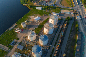 Sunset aerial view of cylindrical storage tanks, pipelines, rail tracks, freight cars, and a road...