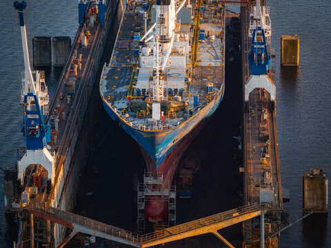 A large ship in a dry dock at Riga shipyard, surrounded by blue and white cranes. The hull features vibrant colors, with scaffolding and equipment visible. - Powered by Adobe