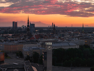 Fototapeta premium Riga's old town spires, including St. Peter's Church and Riga Cathedral, stand against a vibrant orange and pink sunset, with a clock tower in the foreground.
