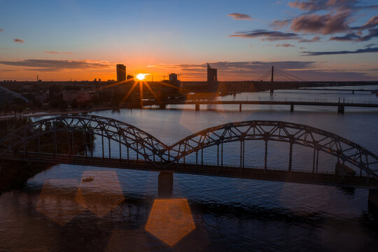 The image shows a sunset over Riga, Latvia, with the Railway Bridge spanning the Daugava River and the Vansu Bridge visible in the background.
