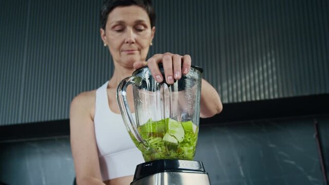 Low angle view of middle-aged Caucasian woman using blender while making fresh green smoothie with organic ingredients at minimalist home kitchen