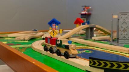 A wooden train engine and car on a model railroad track in a cityscape diorama built on a wooden table with a background of a grey wall. 