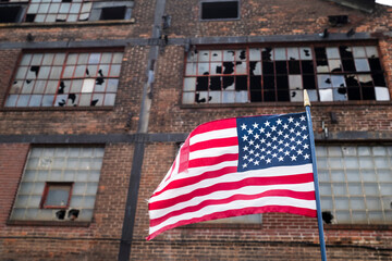American flag waves in front of an abandoned factory with broken windows, symbolizing the decline...