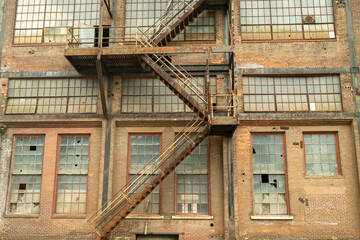 Exposed brick building exterior featuring a rusted metal fire escape and numerous broken windows, illustrating urban decay and abandonment