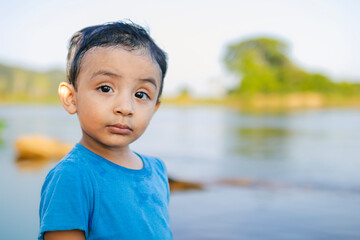 Little boy in a blue shirt looks at the camera by the river during summer vacation outdoors