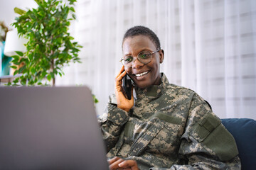Smiling african american female soldier talking on phone and using laptop at home