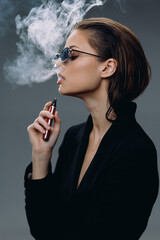 Stylish woman vaping with smoke exhaling, wearing sunglasses in a dark outfit, profile portrait...