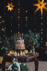 Christmas and New Year beautiful elegant family dinner table setting decorated with candles, fir tree branches, handmade craft toys. Gorgeous big homemade cake with gingerbread houses as centrepiece.