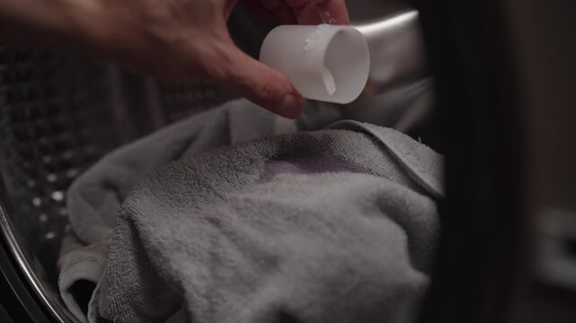 Measuring cup pouring liquid detergent onto stained gray towel inside washing machine, readying clothes for thorough cleaning process. Housewife pouring washing machine detergent from measuring cup.