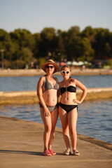 Fototapeta premium Two women in swimwear and hats pose by a body of water on a sunny day. Concept of summer, vacation, relaxation.