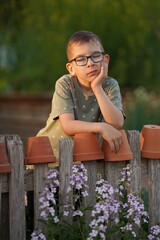 Young boy wearing glasses leans on a fence by purple flowers, surrounded by stacked pots. Concept...