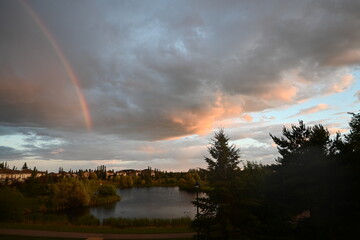 A rainbow sitting above a suburban pond, with a cloudy sky after rain. Taken from a backyard