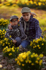Two kids in caps and plaid shirts sit among bright yellow flowers, with one arm around the other....