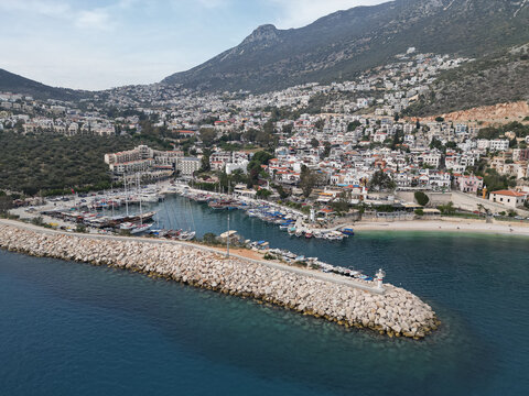 Kas harbor curves below steep hills in Antalya Province, Turkey, with a stone breakwater and small lighthouse, clear turquoise water and clustered white houses, calm midday light and gentle sea breeze - Powered by Adobe