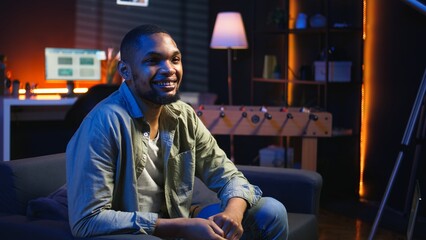 Fototapeta premium Portrait of black freelancer laying on the sofa and texting on phone, relaxing and smiling in his modern loft. Man solving daily teleworking tasks from the living room with cool lights. Camera A.