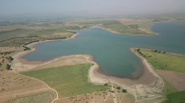 Drone glides over the vast Djizak Reservoir shoreline. Gently curving sandy banks meet clear water in smooth transitions. Fading fields and distant orchards stretch beneath hazy skies as scene unfolds