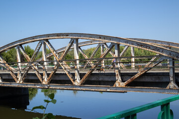a weathered, rusty arched metal bridge spans a tranquil river on a sunny day. its intricate truss structure is reflected in the calm blue water, surrounded by green trees and a clear sky