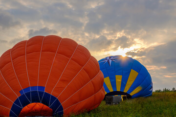 Colorful hot air balloons being inflated at sunrise in a scenic meadow, vibrant orange and blue fabric against dramatic cloudy sky, adventure travel and outdoor festival concept, panoramic photography