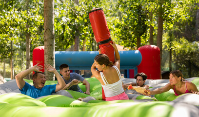 Adults having fun on inflatable amusement playground. Emotional young girl fighting off her friends with inflatable log while they trying to filch toy chickens