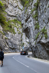 scenic road winds through a narrow canyon, with a car driving away and a person walking in the...