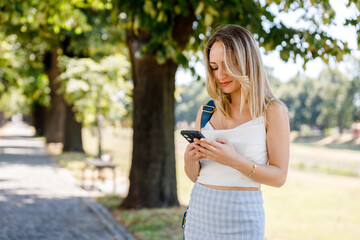 Young woman checks her smartphone while walking in a sunlit park on a warm summer day