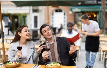 Joyful husband and wife sit on the terrace of a restaurant in warm outerwear and chat, they drink...