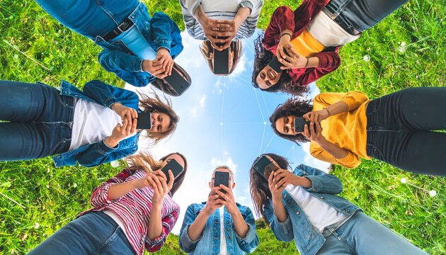 A diverse group of young people stand in a circle outdoors, each engrossed in their smartphones, with a bright blue sky and green grass visible.