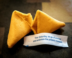 Two golden fortune cookies rest on a dark surface, one broken open to reveal a slip of paper with...
