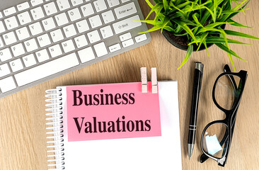 Notepad with business valuations text on pink paper with keyboard, pen, eyeglasses and plant