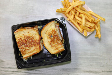 A grilled chicken sandwich on a sourdough bread on a disposable to go container and view of French...