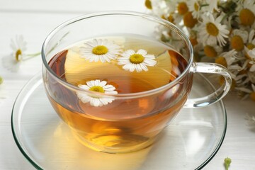 Aromatic tea in glass cup and chamomile flowers on white table, closeup