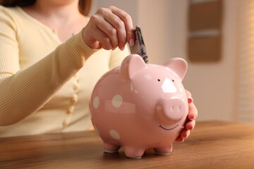 Woman putting money into piggybank at wooden table, closeup