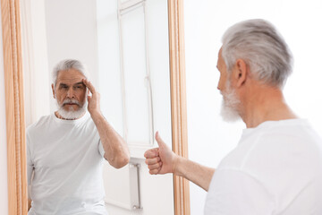 Senior man showing thumbs up in front of mirror while his reflection demonstrating thoughtful face...