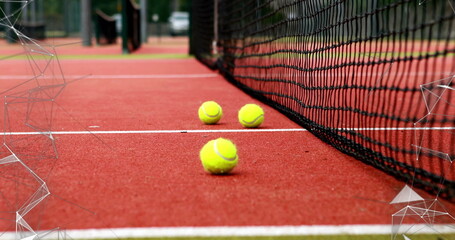 Three yellow tennis balls resting on red clay court near net, with chain-link fence, trolley wheel