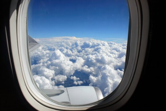 High-altitude view of fluffy white clouds from an airplane window.  Vast expanse of cumulus clouds beneath a clear blue sky.  Airplane window - Powered by Adobe