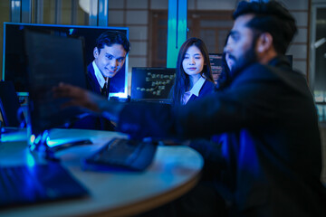 A diverse team of AI developers in a late-night coding session. A senior engineer uses a tablet to guide a programmer as they collaborate on building a complex neural network.