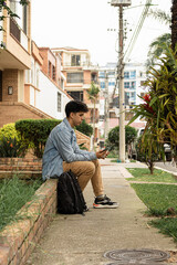 Young man in a denim jacket holding a mug and checking his phone in a quiet urban garden. Colombian...