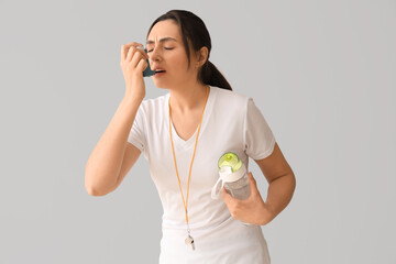 Sporty young woman with water bottle using inhaler on light background