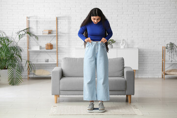 Beautiful young shocked sporty woman in loose jeans with scales at home. Weight loss concept