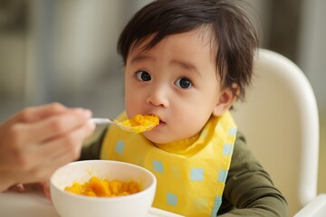 Adorable baby eating pureed pumpkin with a yellow bib, being fed by an adult. Concept for healthy baby food, family nutrition, and parental care