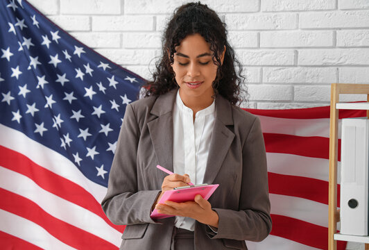 Beautiful young happy African-American businesswoman with USA flag and clipboard in office - Powered by Adobe