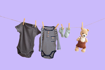 Baby bodysuits with socks and toy hanging on rope against lilac background