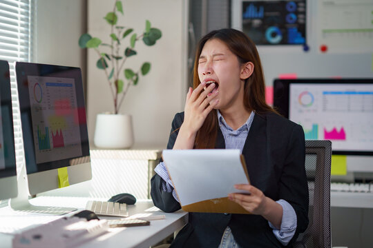 Tired young Asian businesswoman yawning while reading a document sitting at her desk in a modern office - Powered by Adobe