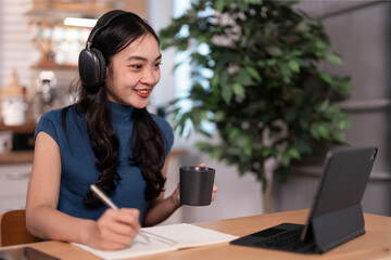 A smiling young Asian woman wearing headphones and holding a mug, is working on a laptop at a desk...