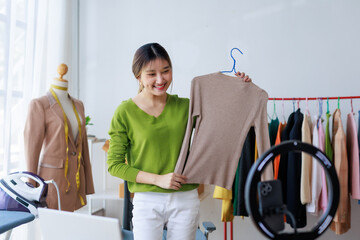 Smiling Asian female designer showing clothes on hanger while recording video for online store in...