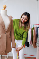 Asian female tailor measuring the sleeve of a suit jacket on a mannequin in her stylish atelier,...