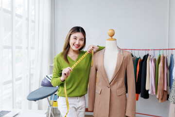 Smiling Asian fashion designer measuring a suit jacket on a mannequin in her stylish atelier,...