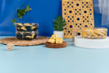 Traditional Indonesian cookies consisting of pineapple-filled nastar and cheese kastengel in clear plastic jars, arranged on woven and marble coasters with a pastel blue background and decorative prop