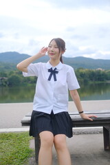 A woman in a school uniform sits on a bench by a lake. A Student girl concept