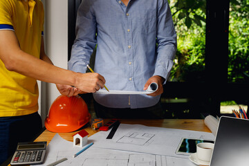 Architects and engineers discussing building blueprint at desk with model house, safety helmet, and...